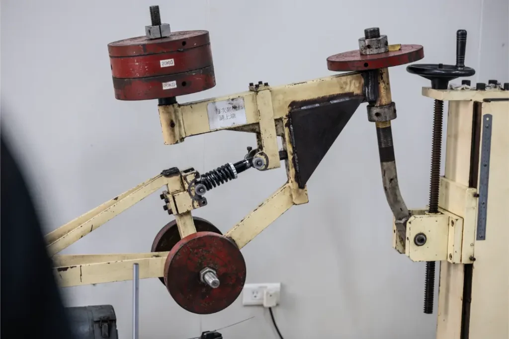 Una máquina de pruebas mecánicas con pesas rojas y un mecanismo de resorte está instalada en el interior, contra una pared blanca con una toma de corriente visible, junto a una tija telescópica inalámbrica RAGE Circuit.
