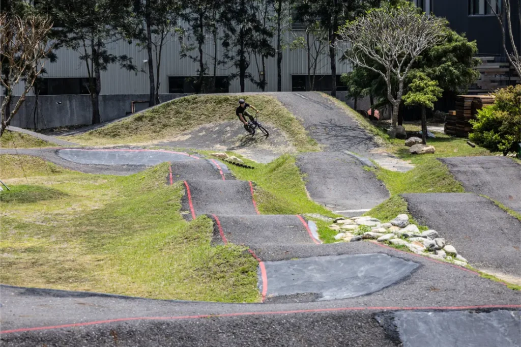 Un ciclista recorre una pista de pump track pavimentada con múltiples baches y curvas, equipada con una tija telescópica inalámbrica, rodeado de césped, árboles y un edificio al fondo.