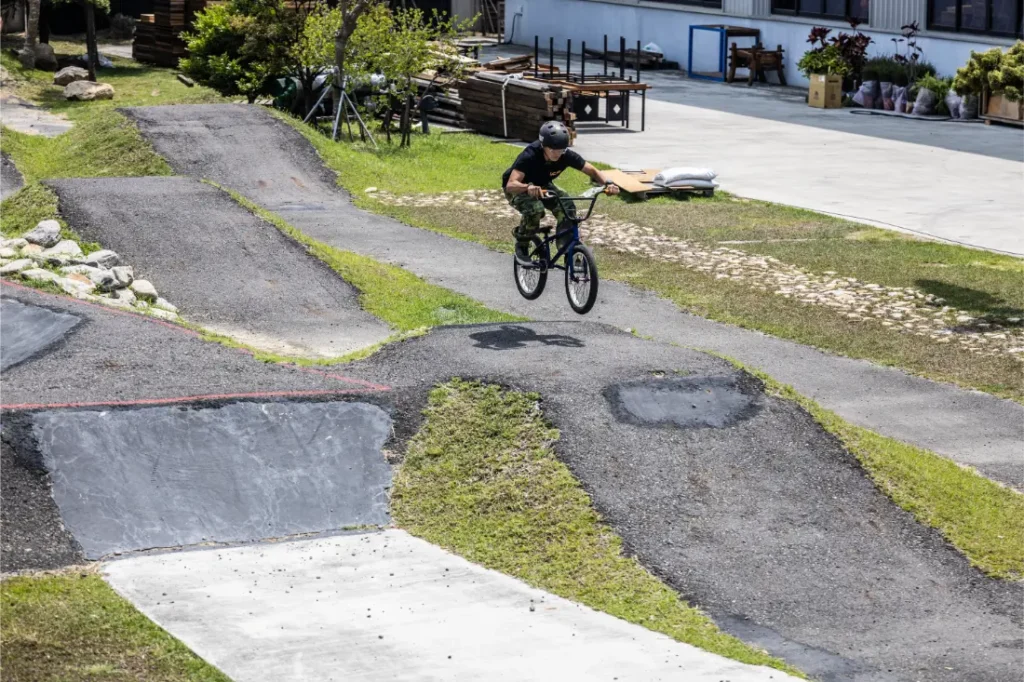 Una persona con casco monta en una bicicleta BMX con tija telescópica inalámbrica RAGE Circuit, cogiendo aire sobre un salto de tierra y asfalto en una pista de pump track al aire libre.