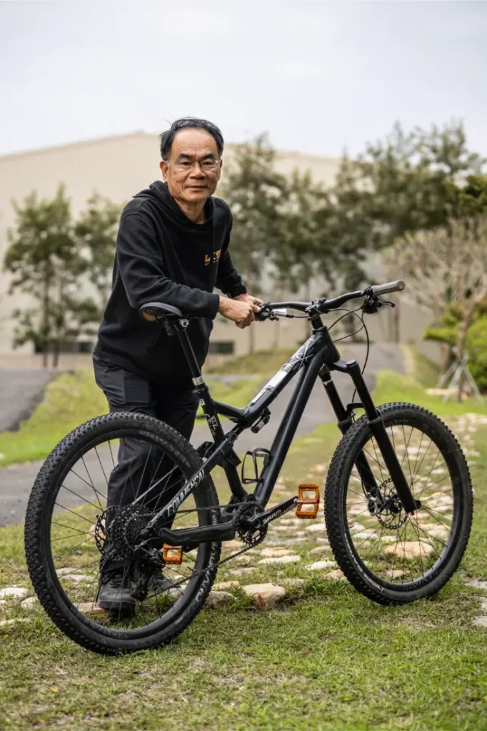 Un hombre vestido de negro está de pie al aire libre sobre la hierba, sujetando una bicicleta de montaña negra con pedales naranjas y una tija telescópica inalámbrica RAGE Circuit. Al fondo se ven árboles y un edificio.