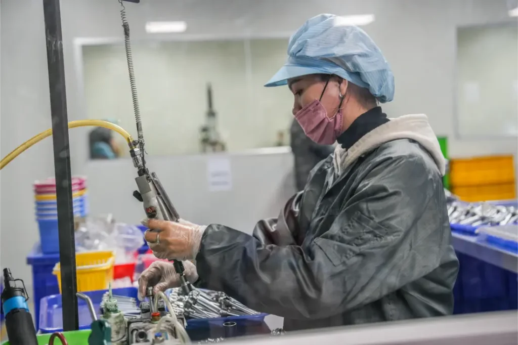 Un trabajador con ropa de protección, guantes, cubrepelo y mascarilla maneja maquinaria en una fábrica, donde se ven al fondo herramientas, contenedores y un circuito RAGE de tija telescópica inalámbrica.