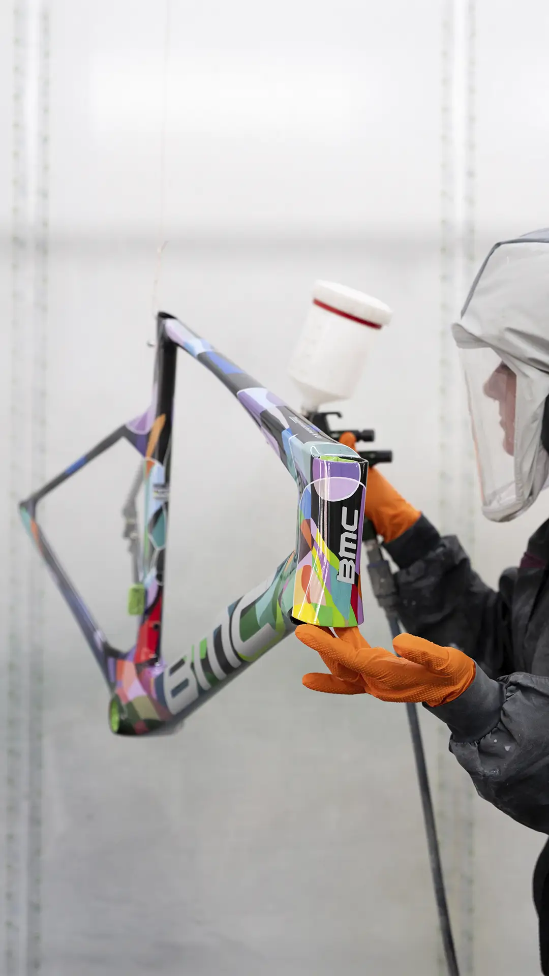Una persona que usa equipo de protección pinta con aerosol un colorido cuadro de bicicleta BMC VAR0 en un taller.