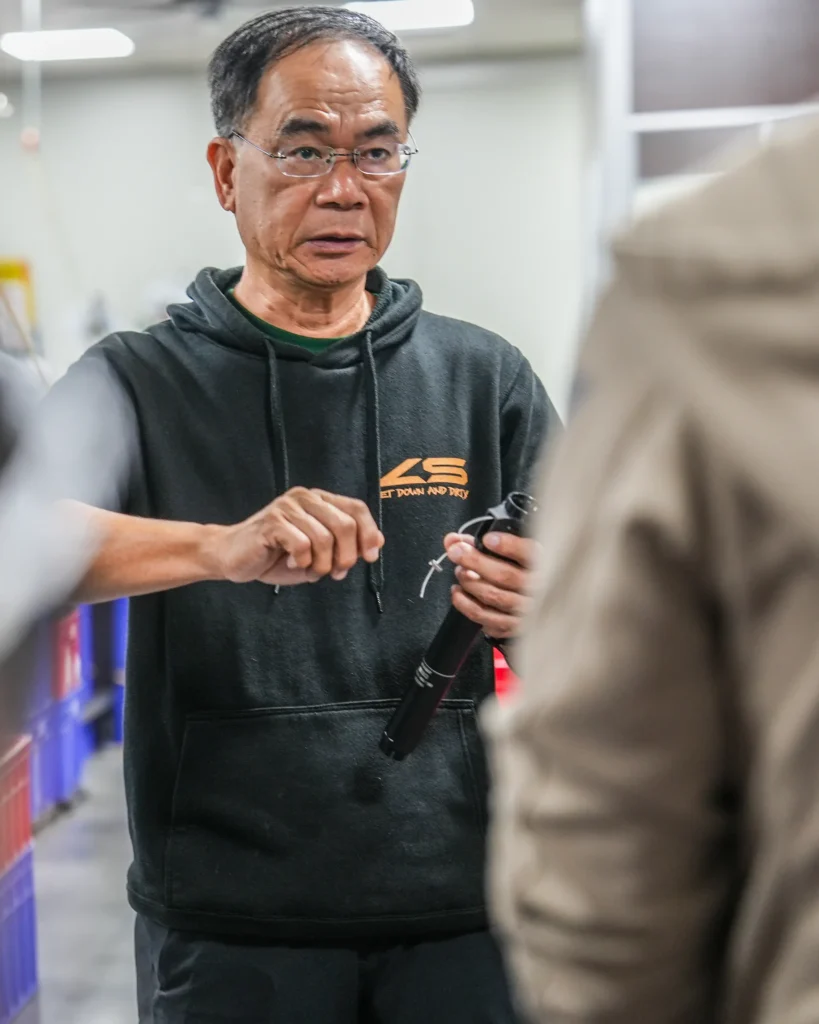Un hombre mayor con gafas y sudadera negra con capucha sostiene una tija telescópica inalámbrica RAGE Circuit mientras habla con alguien en un entorno industrial de interior.
