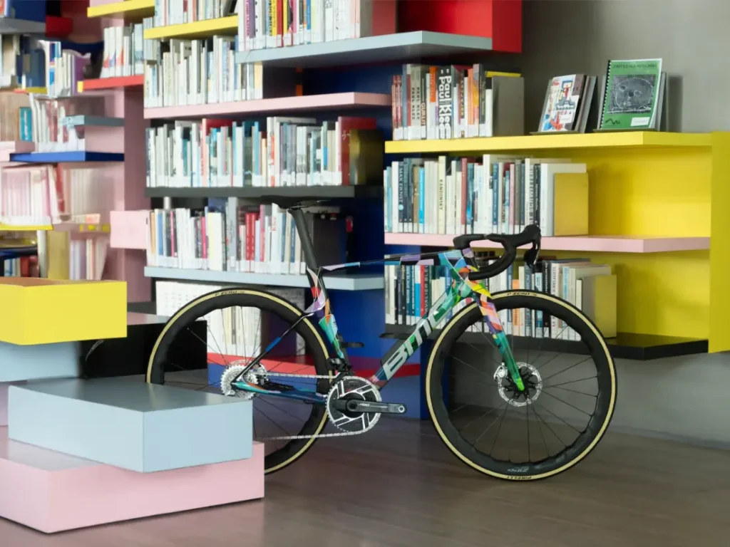 Una colorida bicicleta de carretera BMC VAR0 está estacionada en el interior frente a estanterías con estantes multicolores llenos de libros y revistas.