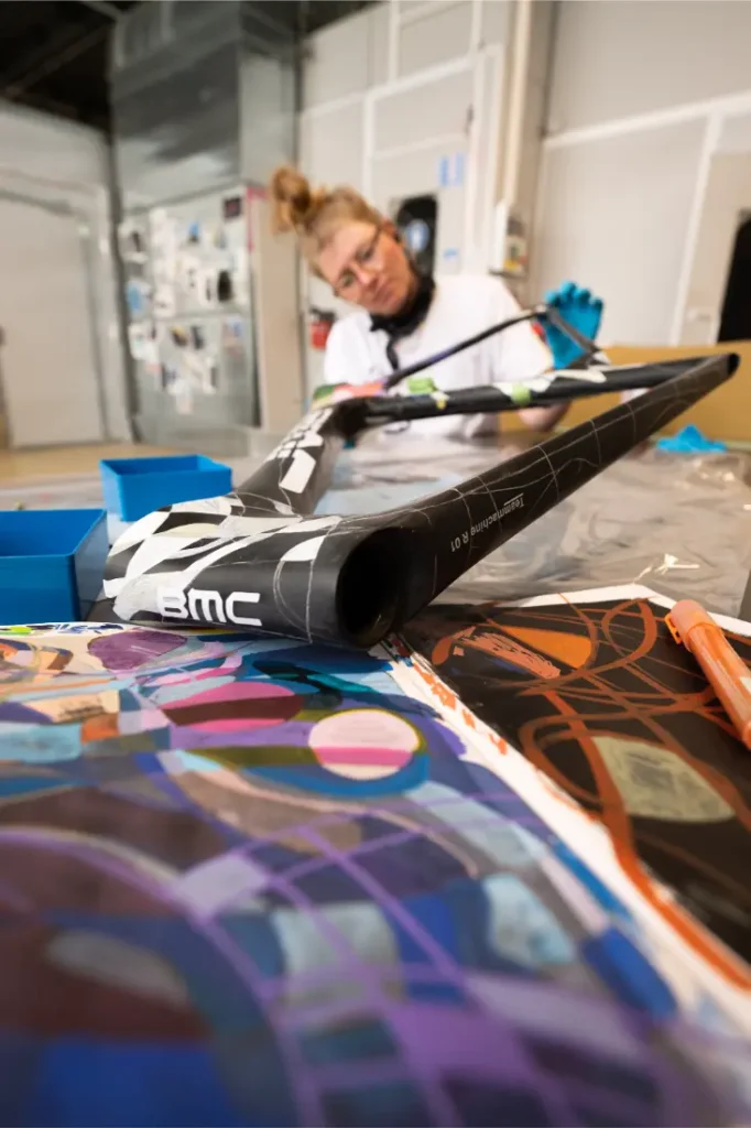 Una persona trabaja pintando un cuadro de bicicleta BMC VAR0 con diseños abstractos coloridos en un taller.