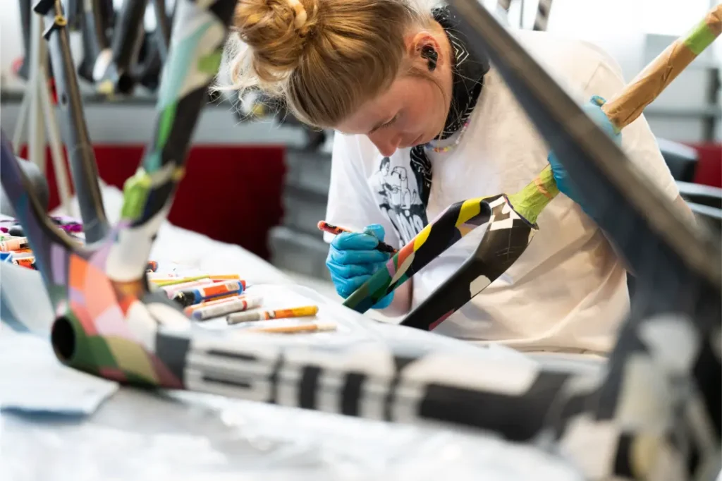 Una persona que usa guantes y tapones para los oídos pinta un patrón colorido en el cuadro de una bicicleta BMC VAR0 usando marcadores, con materiales de arte y otros marcos visibles en el fondo.