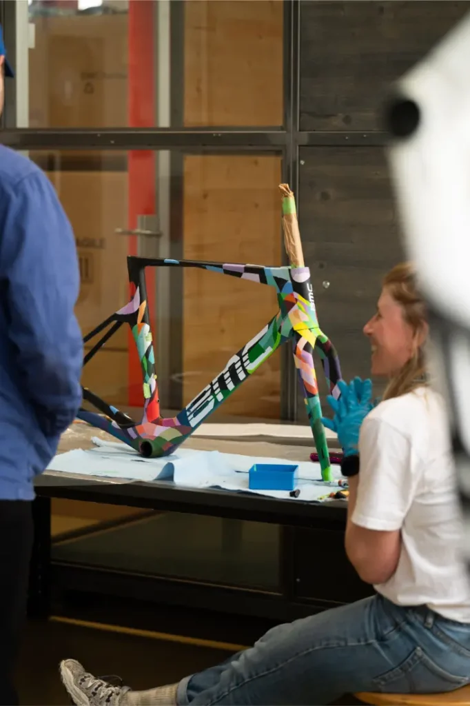 Una persona se sienta sobre una mesa junto a un cuadro de bicicleta BMC VAR0 con un colorido diseño geométrico, mientras otra se encuentra cerca, en un taller.