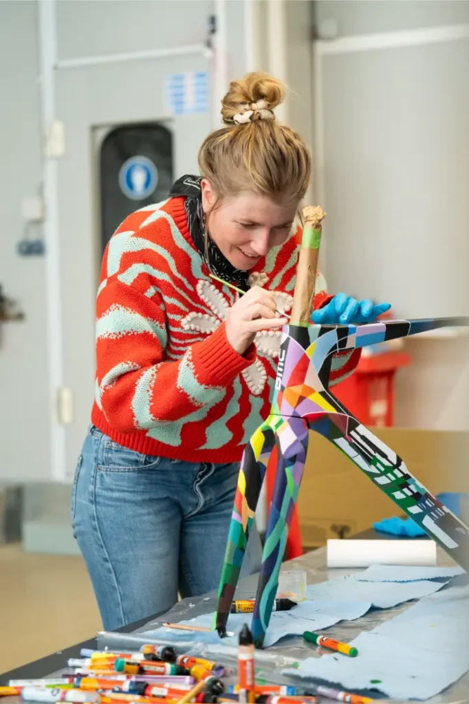 Una mujer que viste un suéter rojo estampado pinta un diseño geométrico colorido en una pequeña escultura de un animal en un espacio de trabajo interior en BMC VAR0.