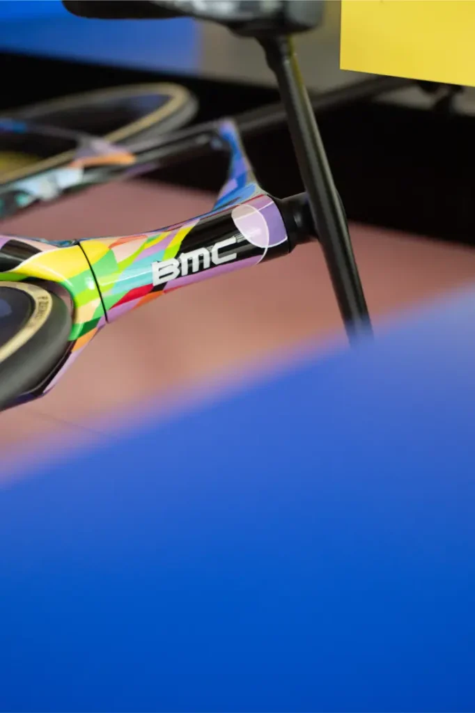 Primer plano de un colorido cuadro de bicicleta BMC VAR0, centrándose en el tubo superior y el poste del asiento, con elementos azules y amarillos borrosos en primer plano y en el fondo.