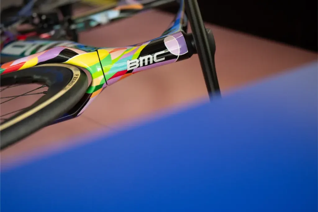 Primer plano de un cuadro de bicicleta BMC VAR0 con un diseño geométrico colorido, centrándose en la horquilla delantera y el neumático, parcialmente oscurecidos por un objeto azul.