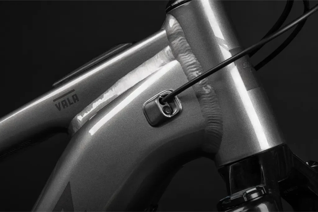 Primer plano de un cuadro de bicicleta metálico de aluminio en el que se aprecian las soldaduras, un orificio para el paso de cables y la marca "Santa Cruz Vala AL" impresa en el tubo.