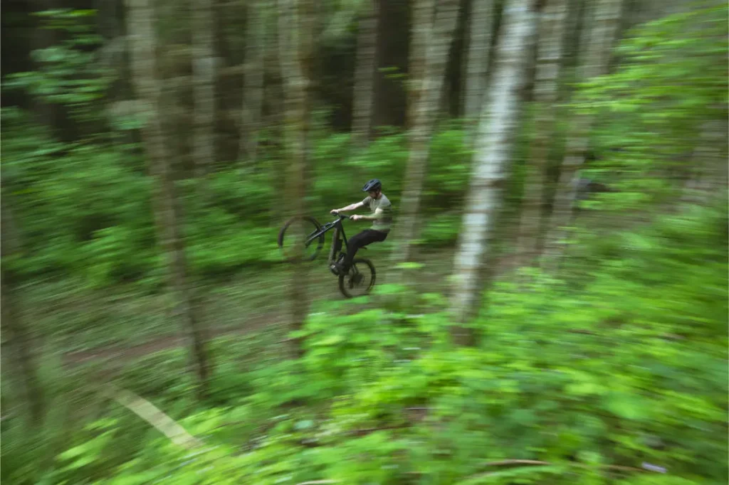 Una persona con casco conduce una bicicleta de montaña Santa Cruz Vala AL por un sendero forestal, levantando la rueda delantera del suelo. El fondo está difuminado con follaje verde y árboles, destacando la agilidad de esta bicicleta con cuadro de aluminio.