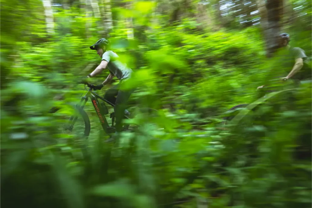 Dos personas montan en bicicletas de montaña Santa Cruz Vala AL a través de un frondoso y verde bosque. La imagen está ligeramente difuminada, lo que resalta la velocidad y agilidad de estos duraderos cuadros de aluminio entre el denso follaje.