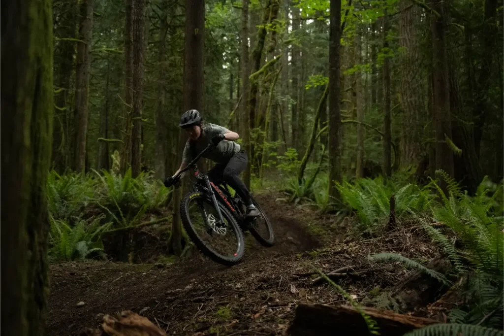 Una persona con casco monta en una bicicleta de montaña Santa Cruz Vala AL de aluminio por un sendero de tierra a través de un bosque denso y verde con helechos y árboles altos.