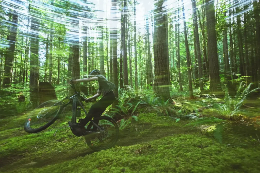 Una persona realiza un caballito en una bicicleta de montaña Santa Cruz Vala AL por un sendero musgoso del bosque, con líneas de movimiento difuminadas que enfatizan la velocidad y el movimiento, resaltando la resistencia ligera de su cuadro de aluminio.