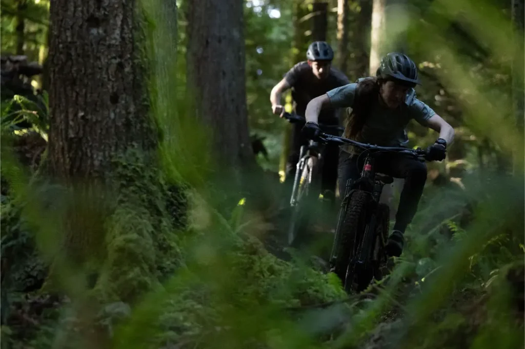 Dos ciclistas con casco montan en bicicletas de montaña Santa Cruz Vala AL, fabricadas en aluminio ligero, por un estrecho sendero boscoso rodeado de altos árboles y denso follaje verde.