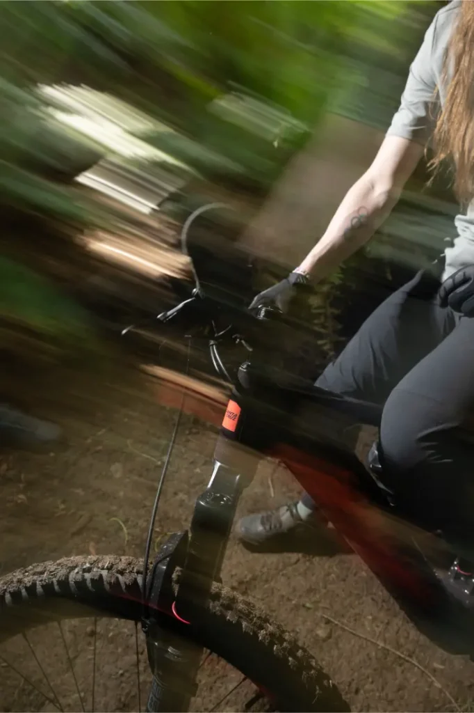 Una persona conduce una bicicleta de montaña Santa Cruz Vala AL por un sendero forestal, con su robusto cuadro de aluminio atravesando los árboles; el desenfoque de movimiento transmite su velocidad, con sólo la parte inferior del cuerpo y un brazo visibles.