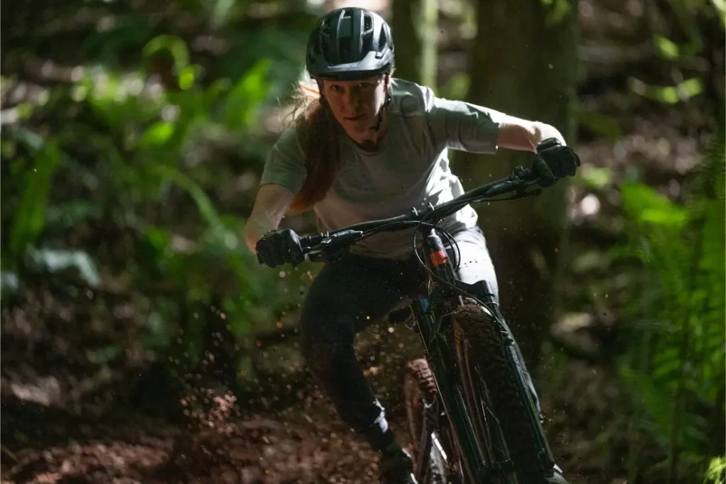 Una persona con casco y guantes monta en una bicicleta de montaña Santa Cruz Vala AL, fabricada en aluminio, por un sendero de tierra a través de un bosque, con la luz del sol filtrándose entre los árboles.
