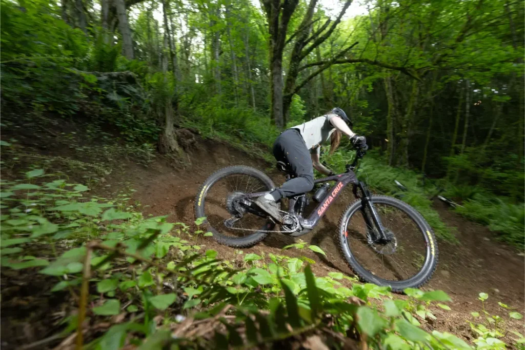 Una persona conduce una bicicleta de montaña Santa Cruz Vala AL con cuadro de aluminio por una curva de un sendero de tierra en un bosque denso y verde.