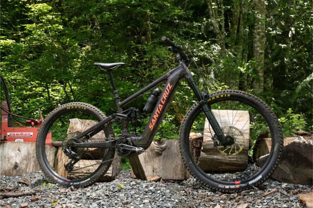 Una bicicleta de montaña negra Santa Cruz Vala AL con gruesos neumáticos y un duradero cuadro de aluminio está aparcada sobre grava delante de tocones de árboles, sobre un exuberante fondo boscoso.
