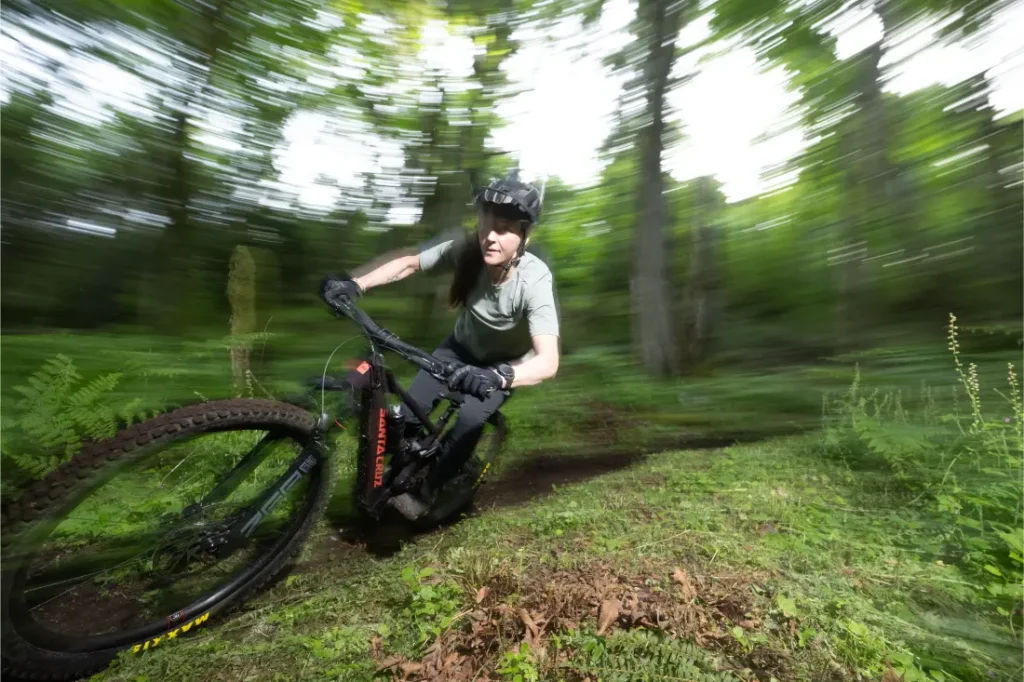 Una persona con casco monta en una bicicleta de montaña Santa Cruz Vala AL, su ligero cuadro de aluminio atraviesa rápidamente un bosque con árboles verdes difuminados al fondo, sugiriendo movimiento.