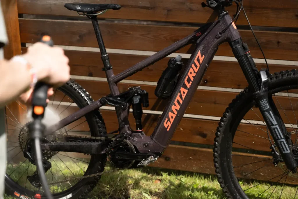Una persona rocía agua para limpiar una bicicleta de montaña negra Santa Cruz Vala AL de aluminio, colocada al aire libre cerca de una valla de madera.