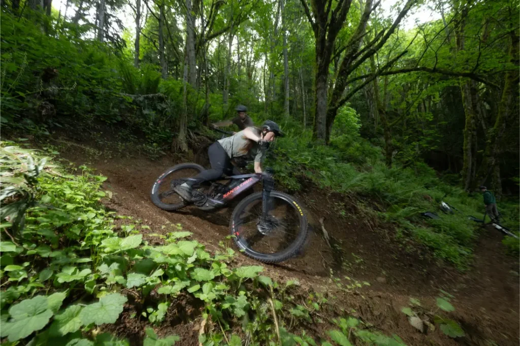 Una persona conduce una bicicleta de montaña Santa Cruz Vala AL con cuadro de aluminio por un sendero de tierra en un bosque denso y verde.