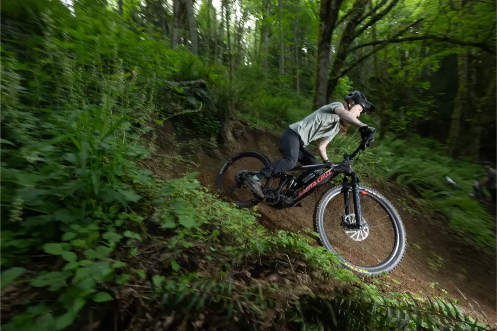 Un ciclista en una bicicleta de montaña Santa Cruz Vala AL desciende a toda velocidad por un sendero forestal, inclinándose en una curva, rodeado de un exuberante follaje verde y árboles imponentes. El ligero cuadro de aluminio garantiza agilidad y control en cada giro.