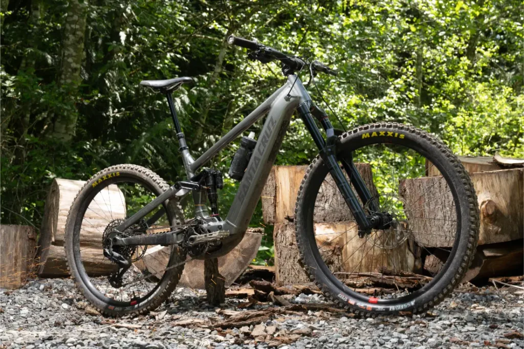 Una bicicleta de montaña Santa Cruz Vala AL con suspensión total, neumáticos anchos y cuadro de aluminio está aparcada sobre grava delante de grandes troncos cortados, rodeada de verde follaje.