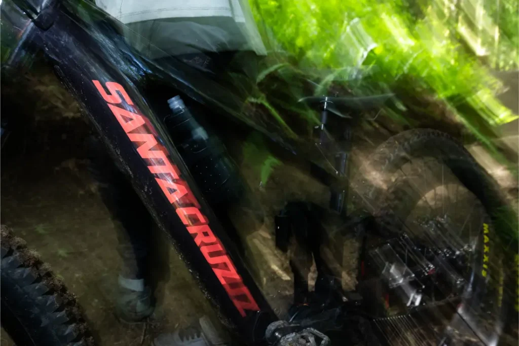 Primer plano de una bicicleta de montaña Santa Cruz Vala AL negra, fabricada en aluminio con llamativas letras rojas, captada con desenfoque de movimiento mientras avanza a toda velocidad por un sendero boscoso.
