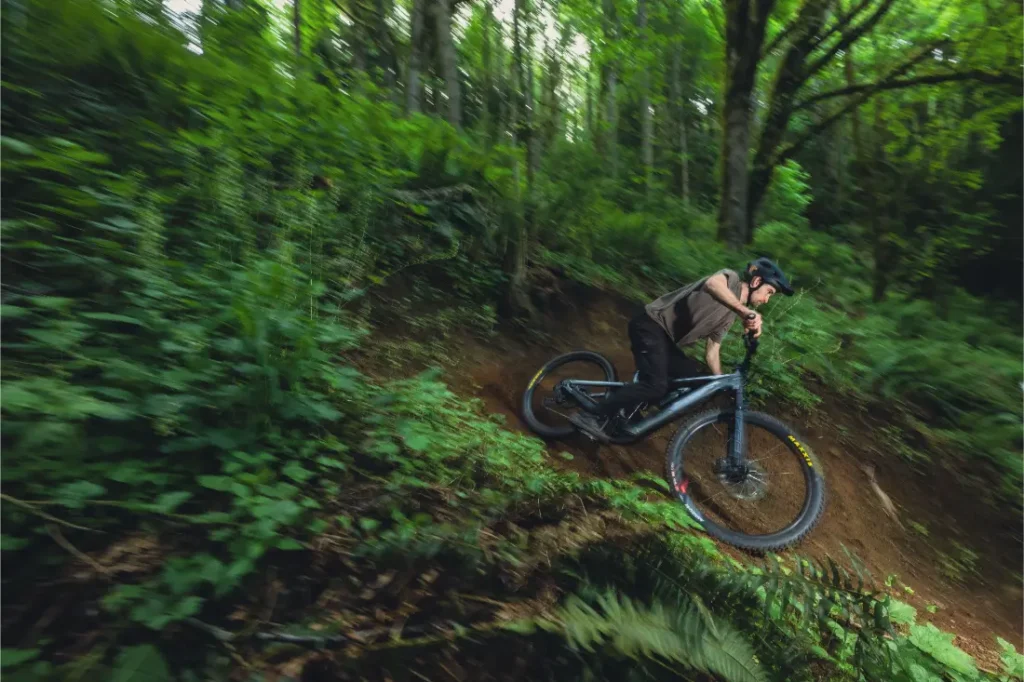 Una persona con casco conduce una bicicleta de montaña Santa Cruz Vala AL con cuadro ligero de aluminio por un sendero de tierra a través de un bosque denso y verde.
