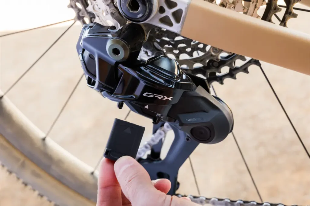 Una mano inserta una pila en el cambio trasero Shimano GRX de una bicicleta, con el casete y la cadena visibles al fondo.