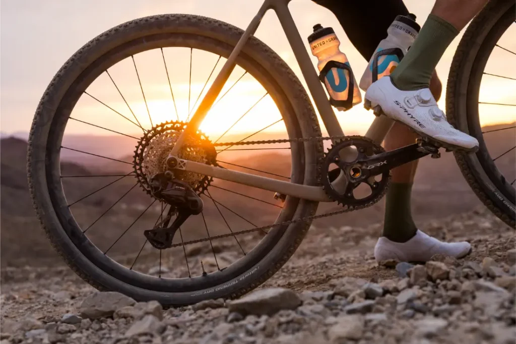Primer plano de las piernas de un ciclista junto a una bicicleta en terreno rocoso al atardecer, con botellas de agua montadas en el cuadro.