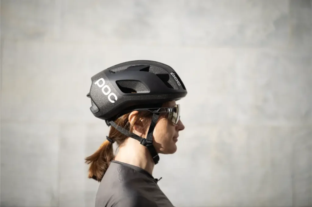 Una mujer de perfil lleva un casco ligero POC Cytal Lite negro y gafas de sol, con el pelo recogido, sobre un fondo liso de color claro, perfecto para circular con calor extremo.