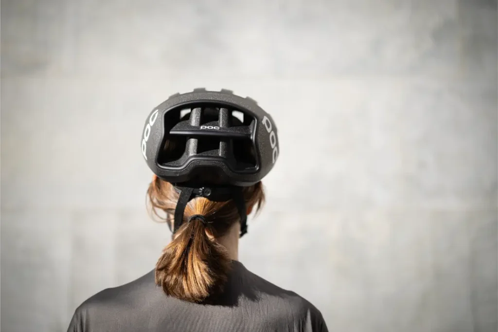 Una persona con el pelo castaño recogido en una coleta aparece de espaldas, con un casco de bicicleta negro ventilado sobre un fondo claro liso.