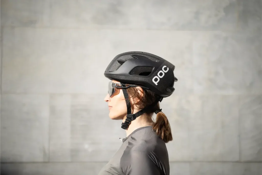 Vista de perfil de una persona que lleva un casco de ciclista POC negro casco ligero, gafas de sol y una camiseta gris, de pie delante de un muro de piedra gris claro.