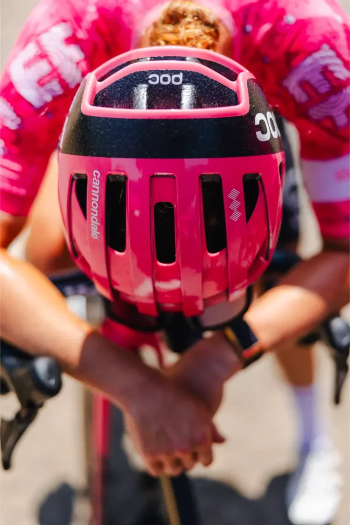 Un ciclista con casco y maillot rosa se inclina hacia delante sobre el manillar, con las manos entrelazadas en posición de carrera, manteniéndose fresco con un casco ventilado durante el calor extremo.