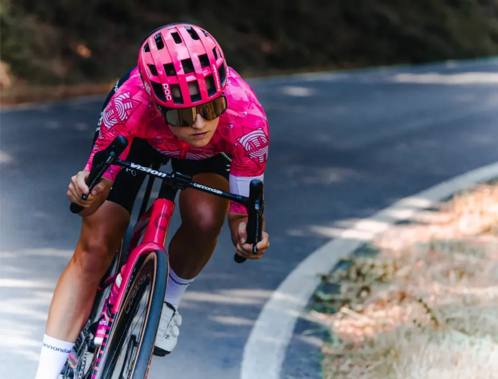 Un ciclista con una equipación rosa y casco ventilado monta en una bicicleta de carretera rosa, inclinándose en una curva de una carretera asfaltada rodeada de árboles y hierba seca.