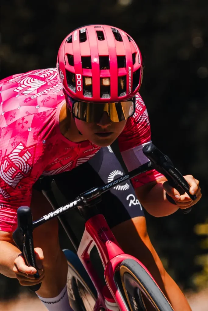 Un ciclista con casco ventilado rosa y maillot a juego monta en bicicleta al aire libre, inclinado hacia delante en posición aerodinámica.
