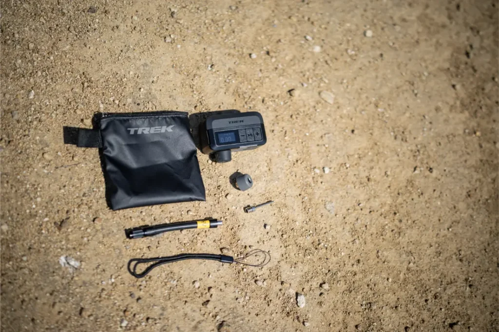 Un manómetro digital para neumáticos, una bolsa TREK negra, un adaptador de válvula, un cordón y una herramienta Trek Air Rush para inflado de ruedas están colocados sobre el suelo arenoso.