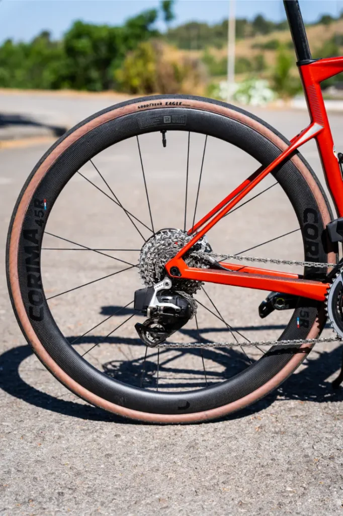 Primer plano de la rueda trasera de una bicicleta roja con llanta de carbono Ruedas Corima 45R, cubierta de pared de color tostado y cassette de cambio visible, que muestra ligereza y rendimiento en una superficie pavimentada al aire libre.