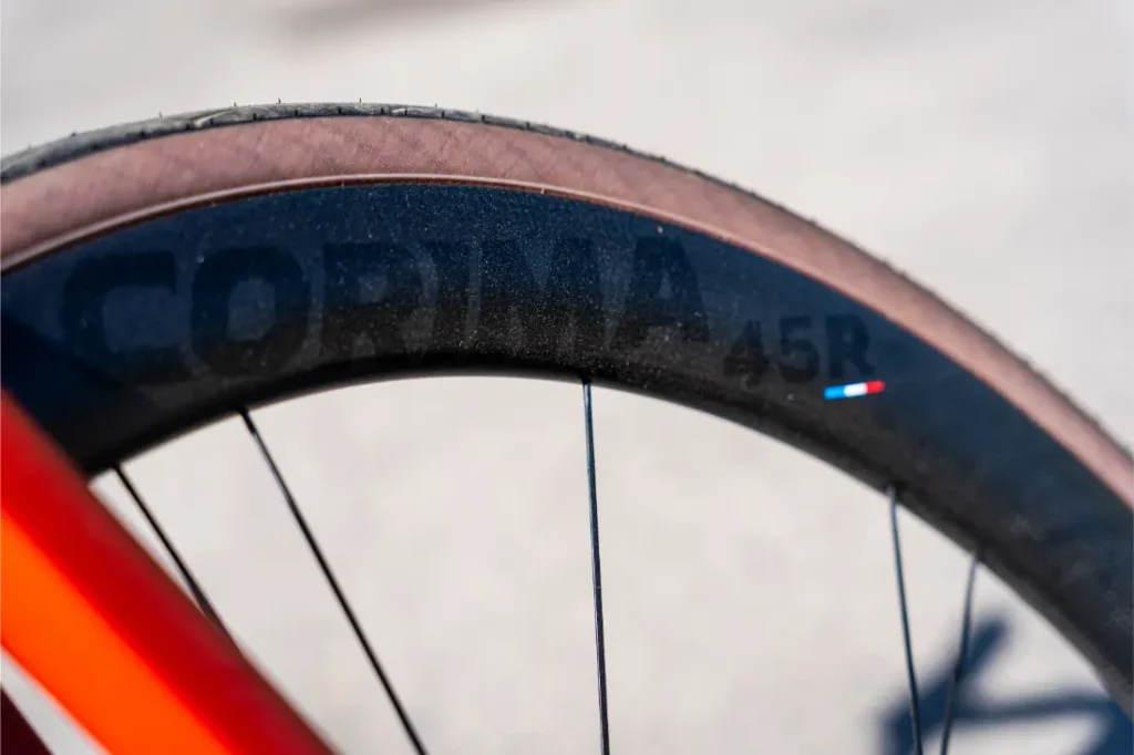 Primer plano de una rueda de bicicleta en la que destaca la marca Ruedas Corima 45R en la llanta negra, famosa por su ligereza y rendimiento, con parte de un cuadro de bicicleta naranja visible al fondo.