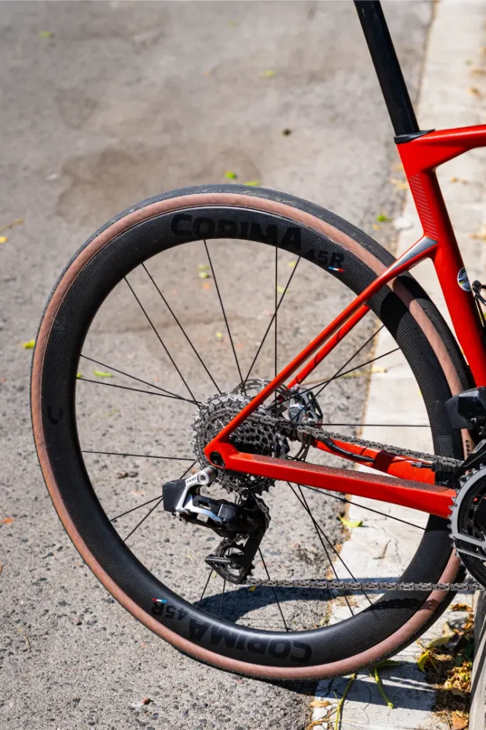Primer plano de la rueda trasera y el sistema de cambios de una bicicleta de carretera roja con Ruedas Corima 45R, aparcada en una calle asfaltada cerca de un bordillo, destacando la ligereza y el rendimiento óptimo.