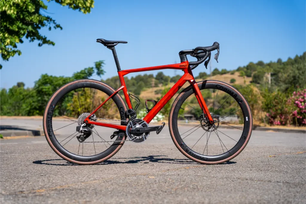 Una bicicleta de carretera roja con Ruedas Corima 45R, manillar rebajado y frenos de disco está aparcada en una superficie pavimentada al aire libre, ofreciendo una ligereza y un rendimiento impresionantes con árboles y colinas de fondo.