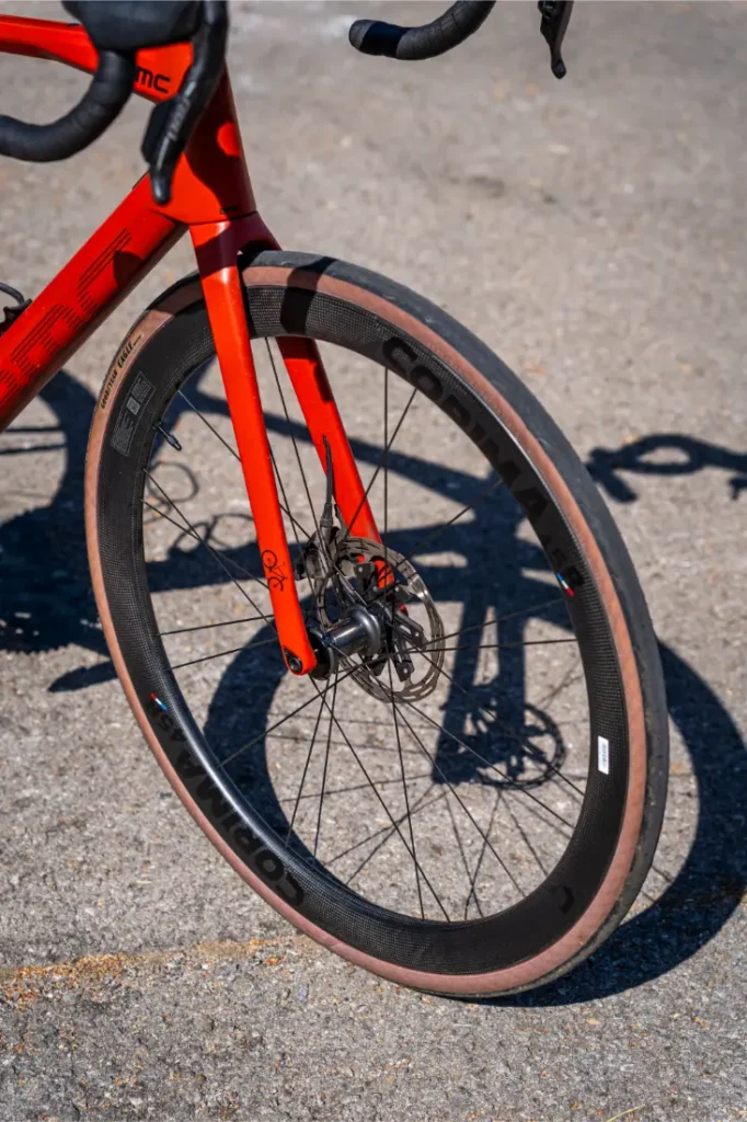 Primer plano de la rueda delantera de una bicicleta de carretera roja con llanta de carbono Ruedas Corima 45R, neumático de flanco marrón, freno de disco y pavimento al fondo, mostrando ligereza y rendimiento.