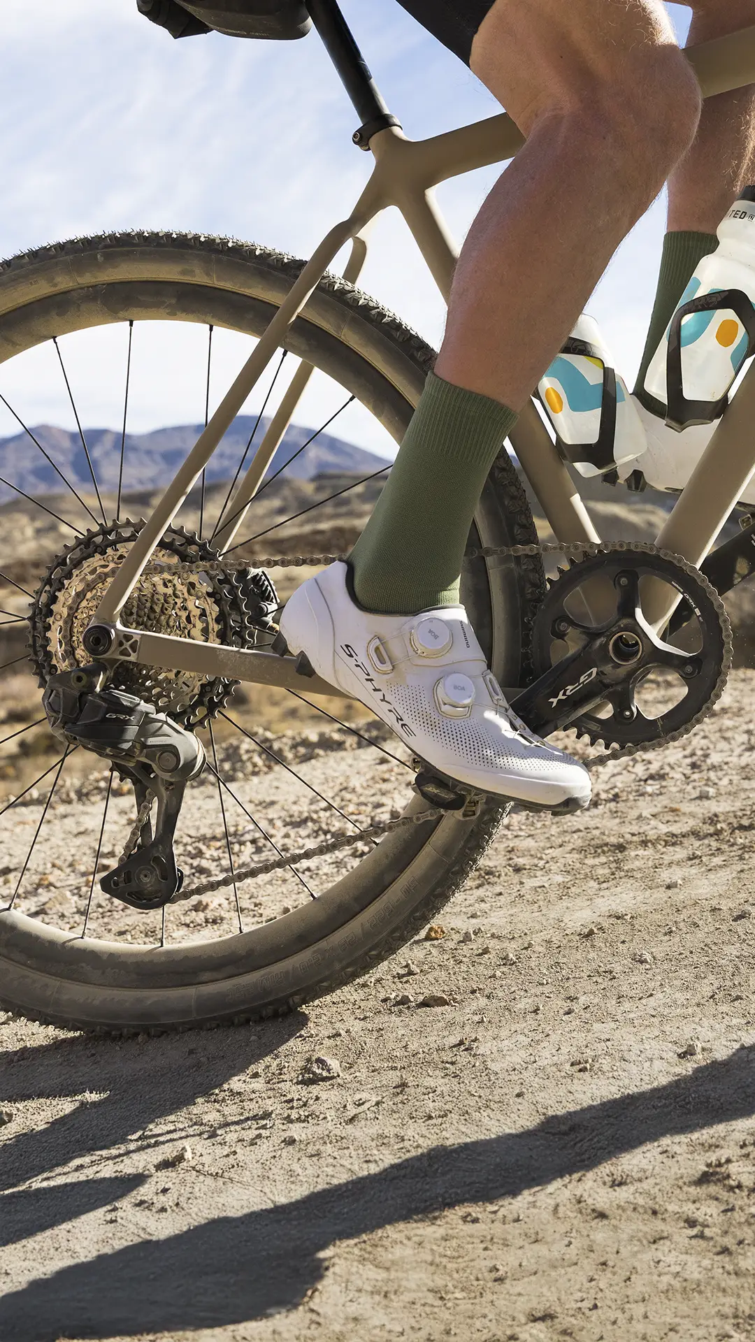 Primer plano de la pierna y la bicicleta de un ciclista en un camino de tierra, mostrando los engranajes de la bicicleta, la biela negra y la zapatilla de ciclismo blanca con calcetines verdes.