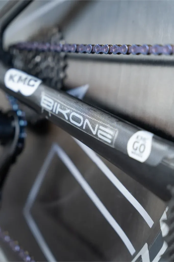 Primer plano de una cadena y un cuadro de bicicleta, mostrando la marca "BIKONE" con una etiqueta "KMC" y una pegatina en la que se lee "EPIC GO original". Las cadenas KMC impulsan al Caja Rural-Seguros RGA en cada trayecto.