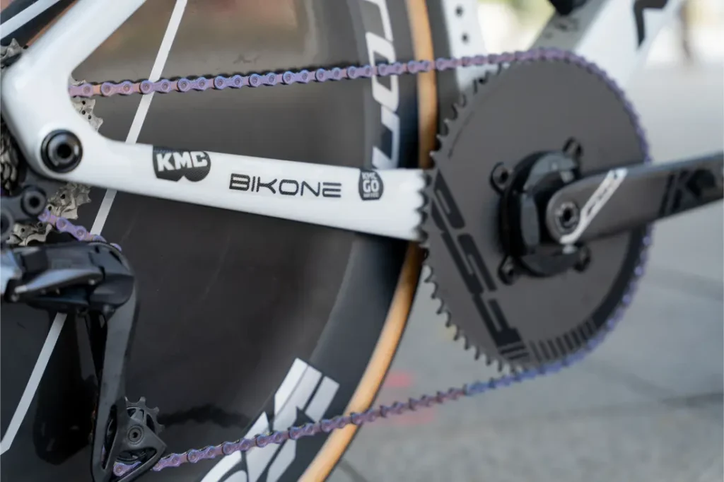 Primer plano de una transmisión de bicicleta con cadena morada, plato grande y bielas de las marcas KMC y BIKONE, como las utilizadas por Caja Rural-Seguros RGA, donde las cadenas KMC impulsan al equipo.