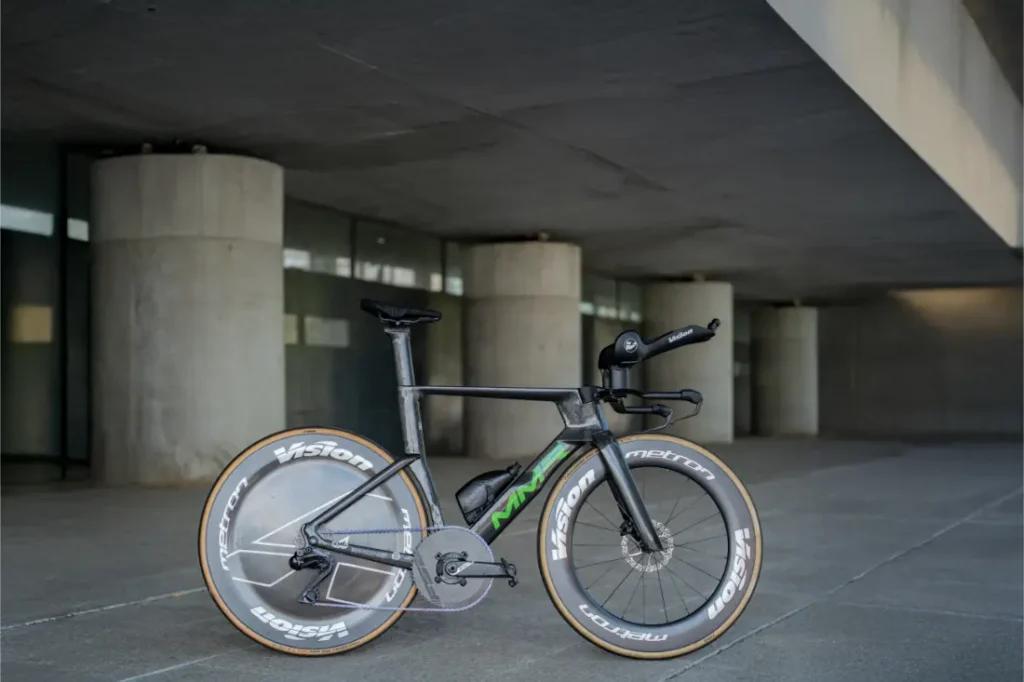 Una moderna bicicleta de contrarreloj con ruedas de disco y de sección profunda, como las que Las cadenas KMC impulsan al Caja Rural-Seguros RGA, está aparcada sobre una superficie de hormigón bajo una estructura volada.
