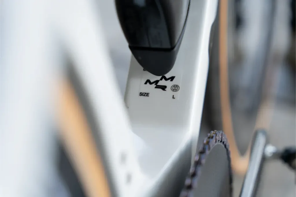 Primer plano de un cuadro de bicicleta blanco que muestra el logotipo MMR y la talla "L" junto a la rueda trasera y el plato. Las cadenas KMC impulsan al Caja Rural-Seguros RGA, destacando la calidad y rendimiento en cada detalle.